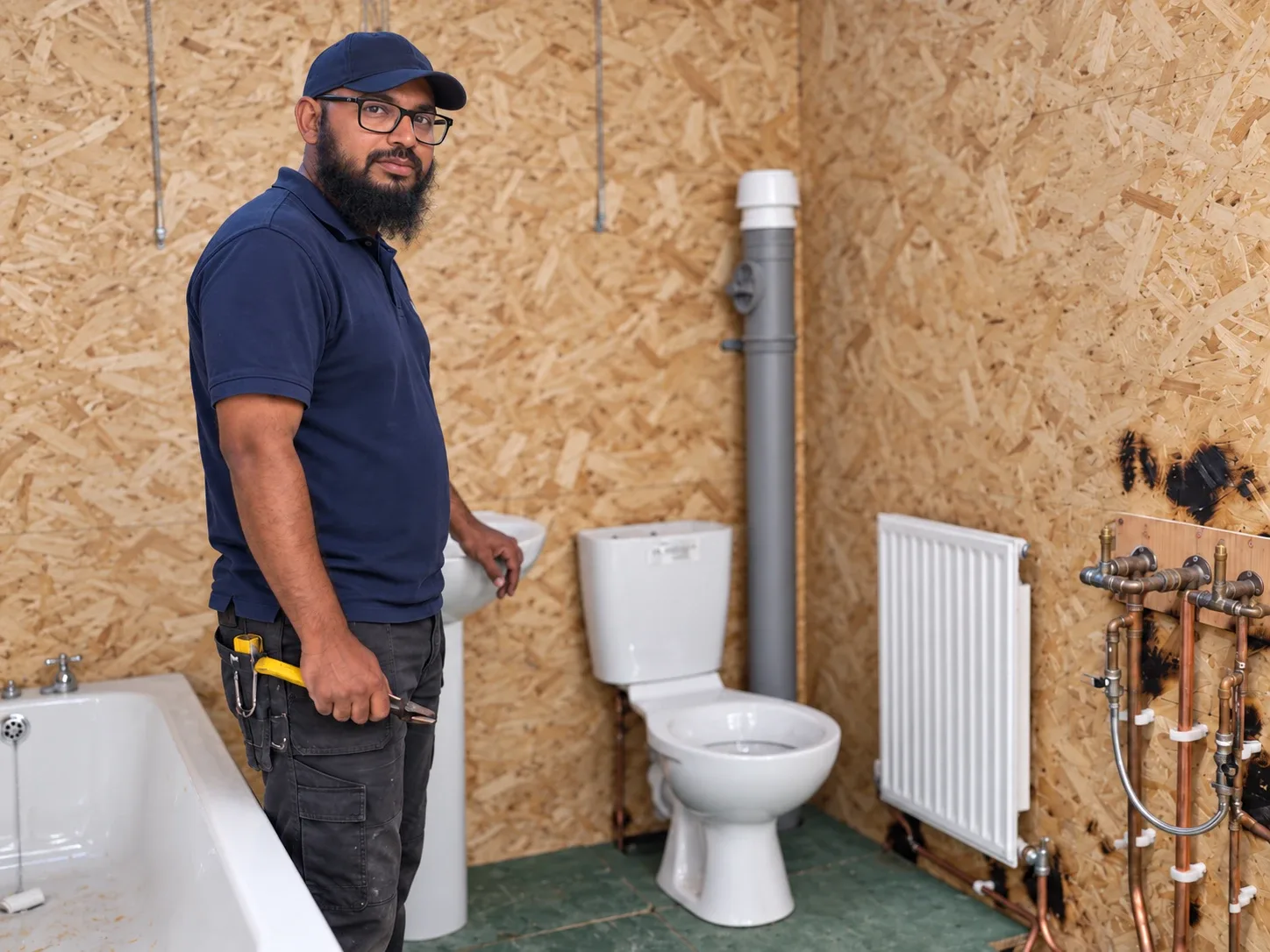 Reduan in a navy polo and cap, standing beside a bathroom suite with pliers in hand